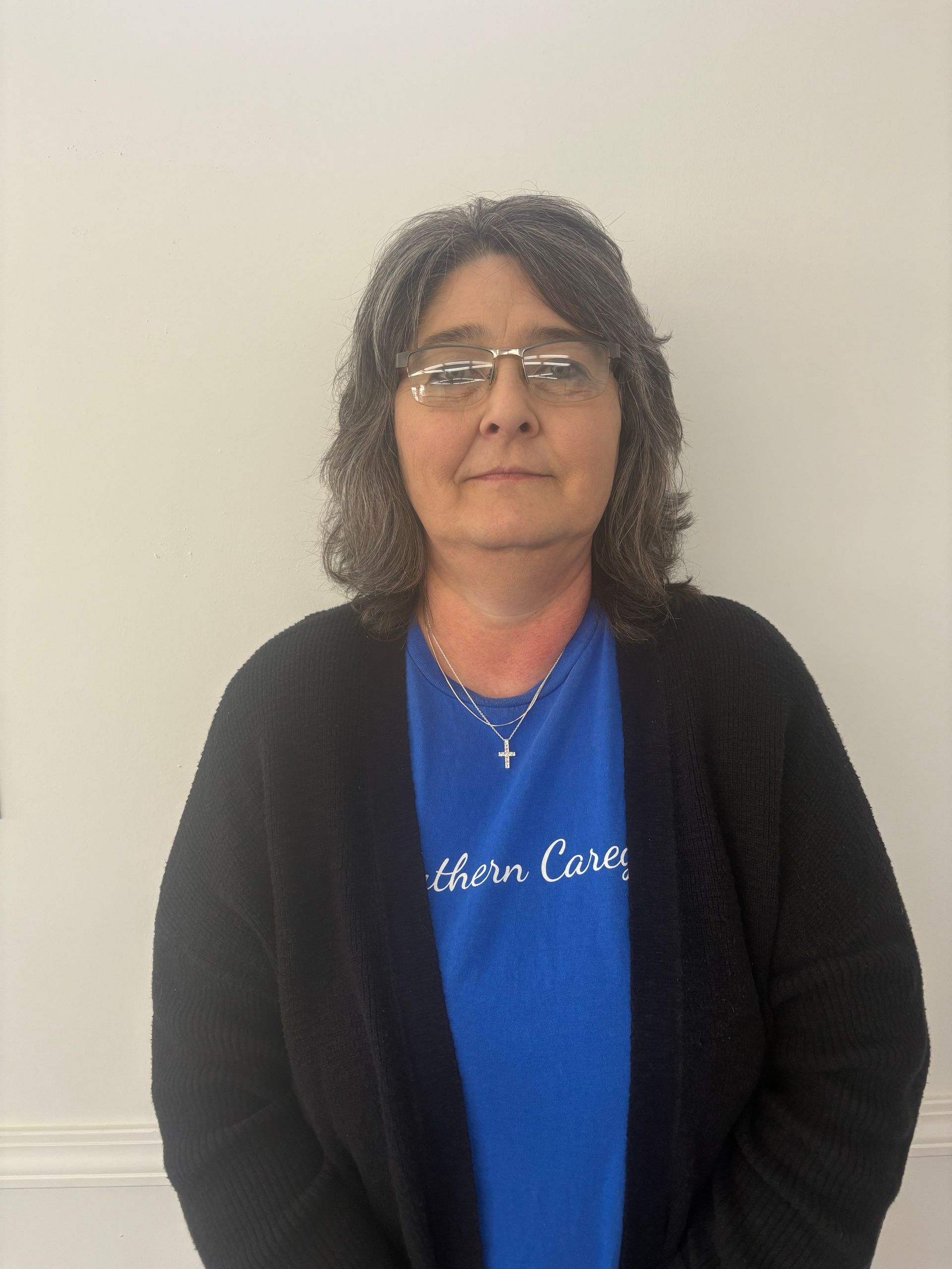 Person standing against a plain wall, wearing glasses, a blue shirt, and a black cardigan.