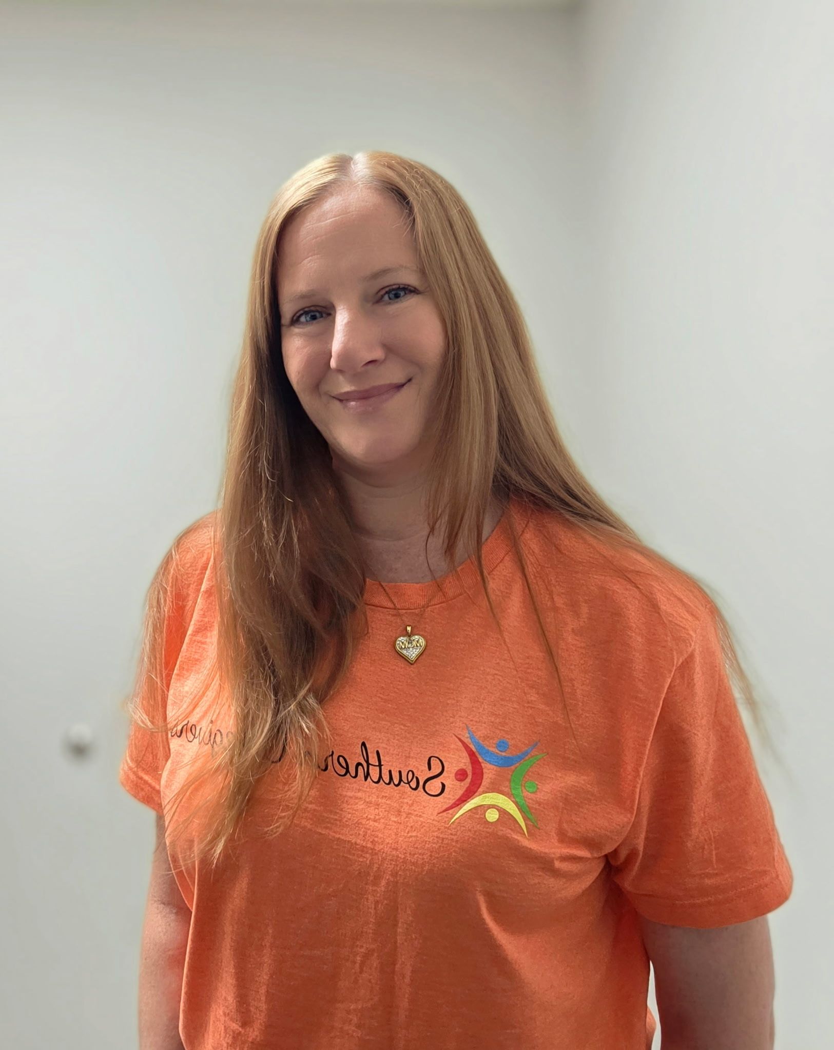 Woman in an orange T-shirt smiling indoors against a plain wall.