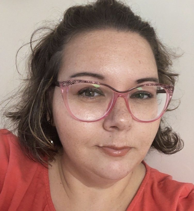 Woman with pink glasses and red shirt, looking at the camera against a plain wall.
