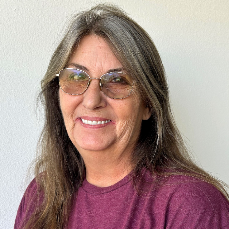 Smiling woman with long gray hair and glasses, wearing a purple shirt against a light wall