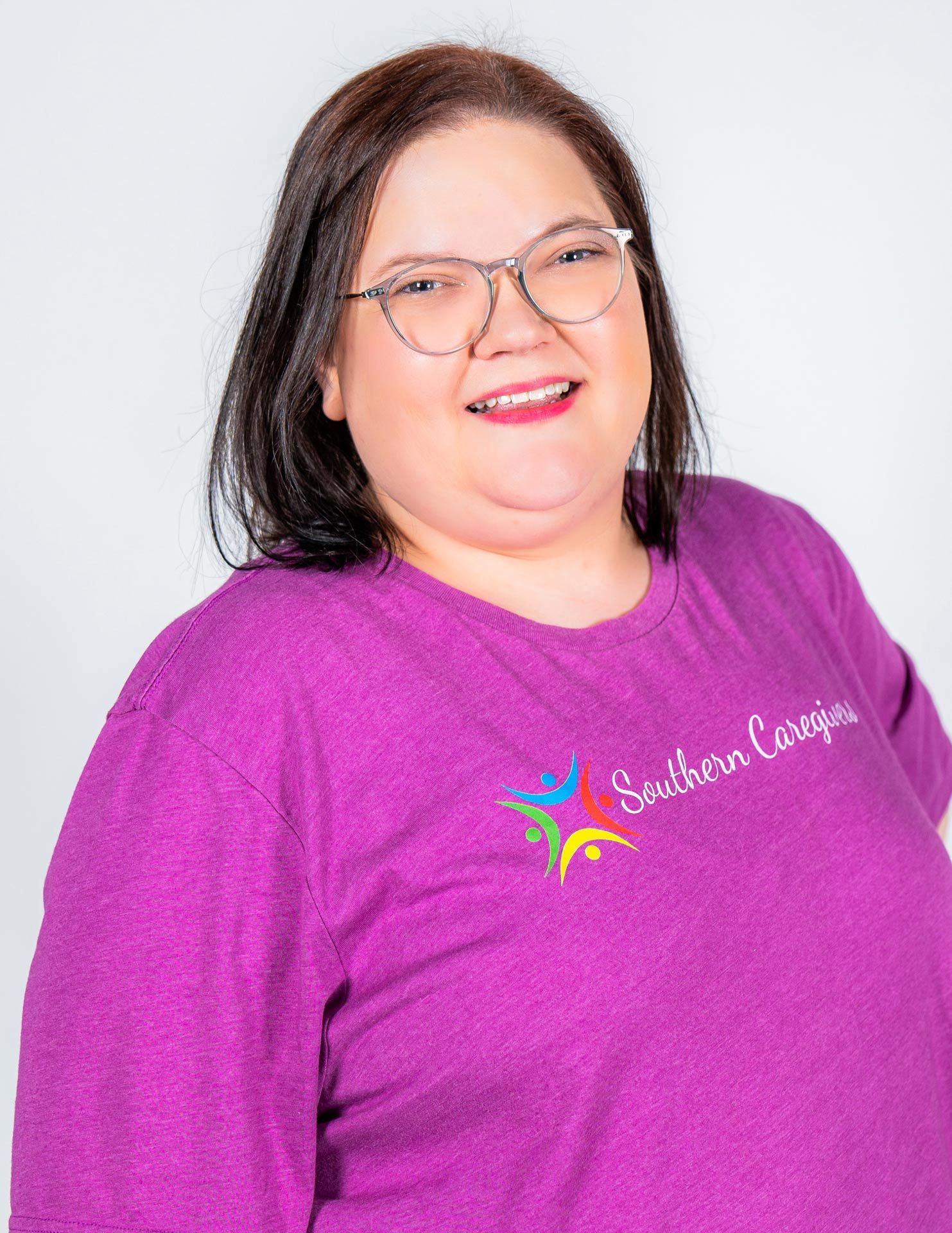Smiling person in glasses wearing a purple shirt with a colorful logo, standing against a light background