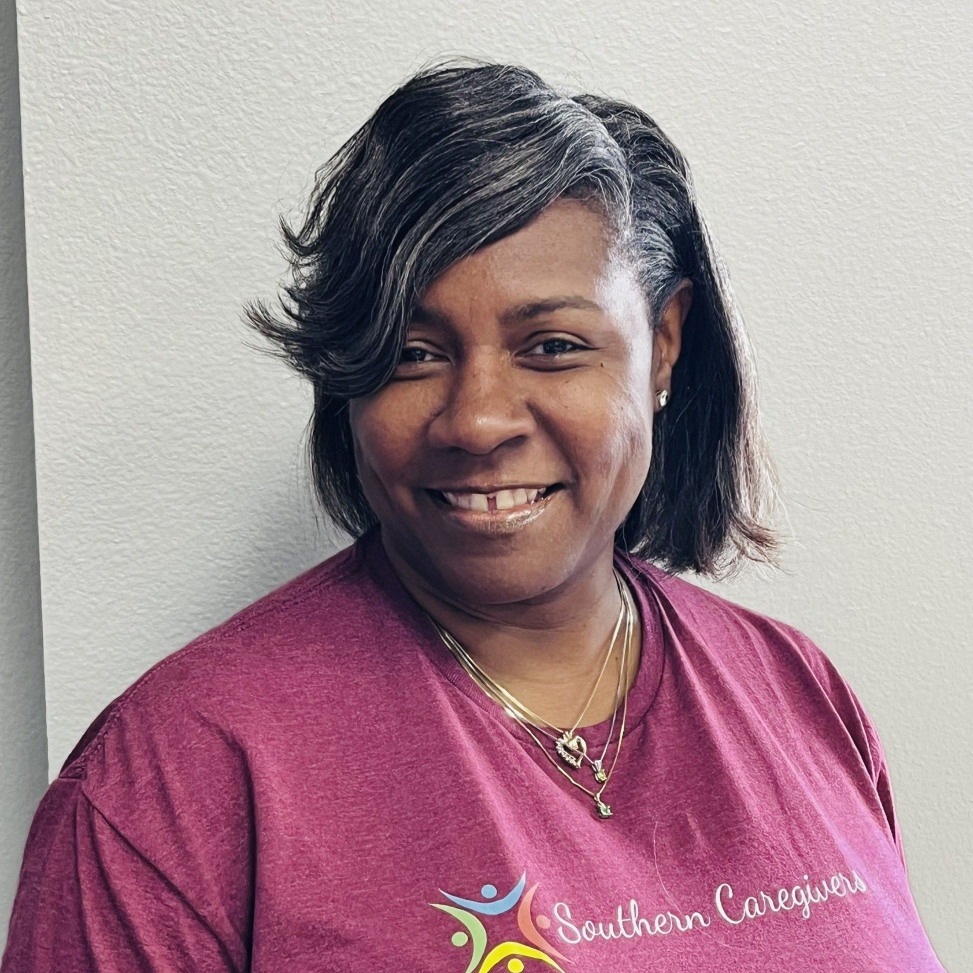 Smiling person in a magenta T-shirt standing against a light gray wall.