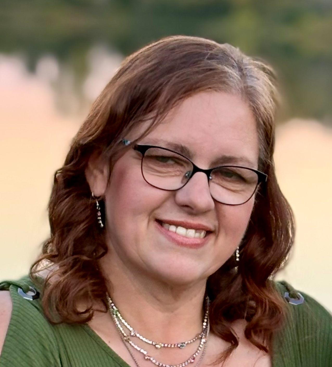 Smiling woman with glasses and wavy brown hair, wearing a green top and silver necklaces outdoors.