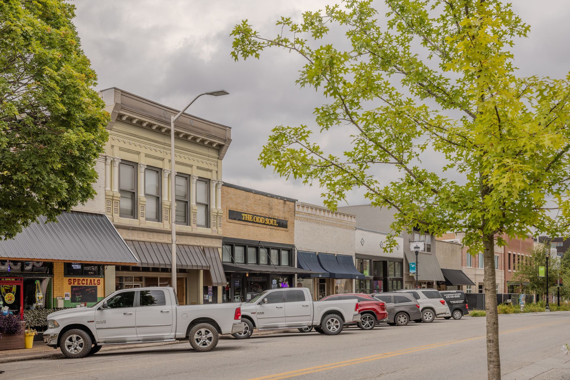Street scene with parked cars in front of low-rise buildings with storefronts under a cloudy sky.