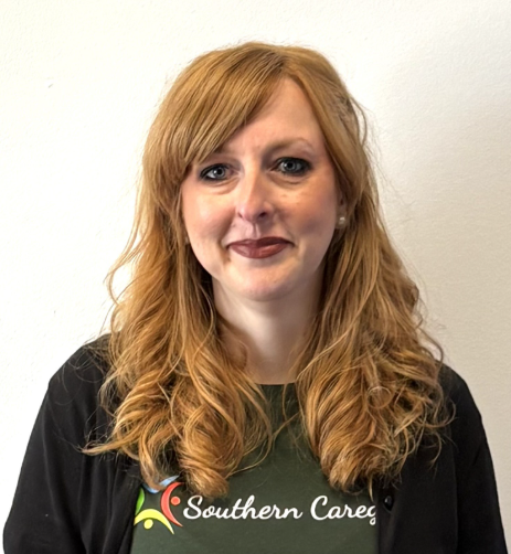 Woman with long red hair wearing a black Southern Care shirt, standing against a white wall.