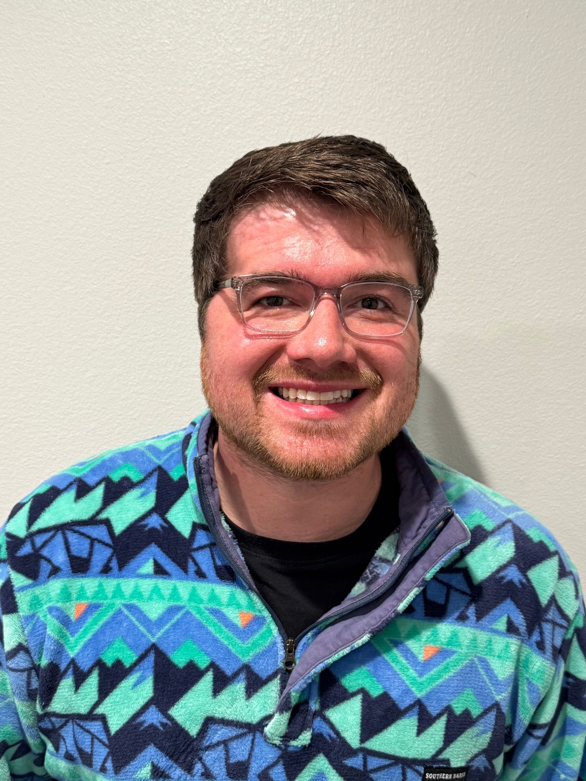 Smiling person wearing glasses and a blue-green patterned sweater against a plain wall