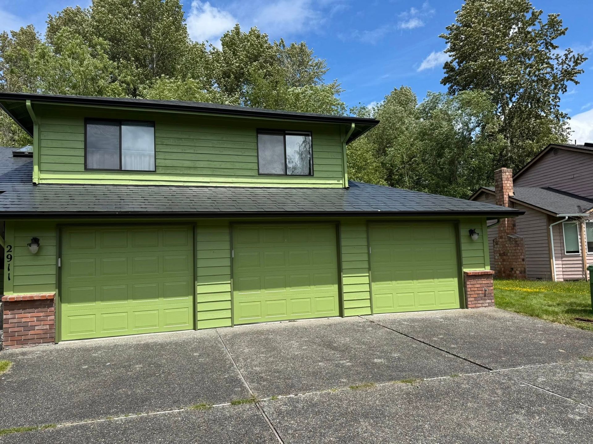 Green house with three garage doors and two upper-level windows. Brick accents, asphalt driveway, and trees.