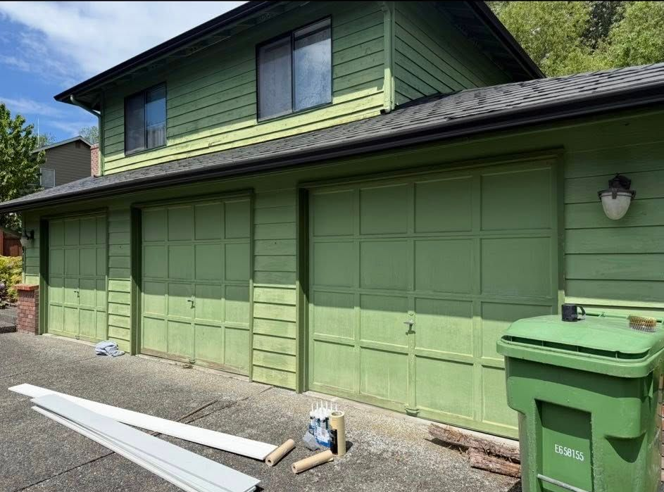 Green house with green garage doors; a green trash can and white trim pieces sit outside.