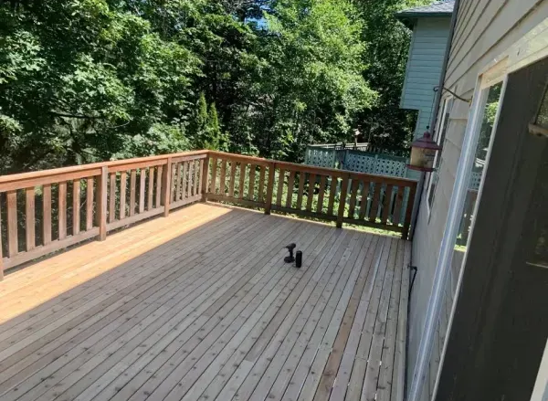 Wooden deck with brown railings, overlooking trees and a small fence. Two dumbbells sit on the deck.