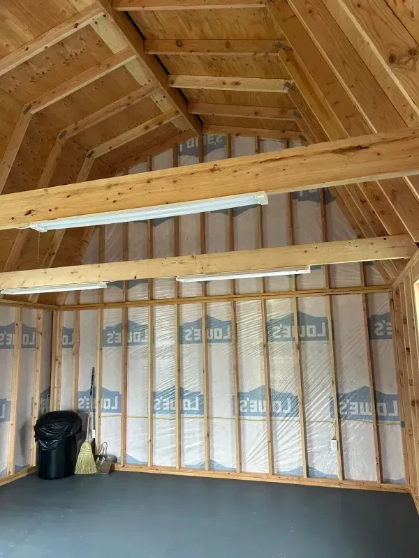 Interior view of a wood-framed building. Insulation lines the walls, exposed beams, and fluorescent lights.