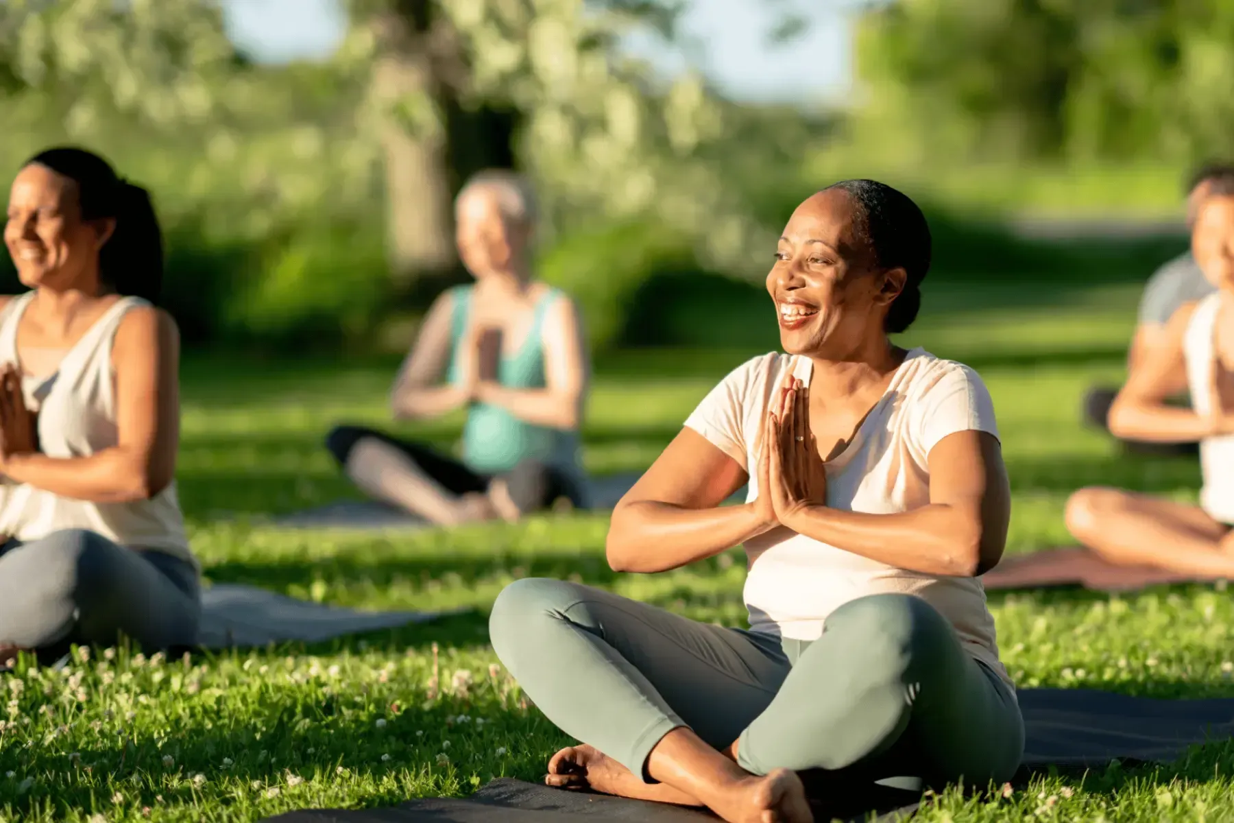 People seated cross-legged outdoors on grass, smiling with hands together in a yoga pose.
