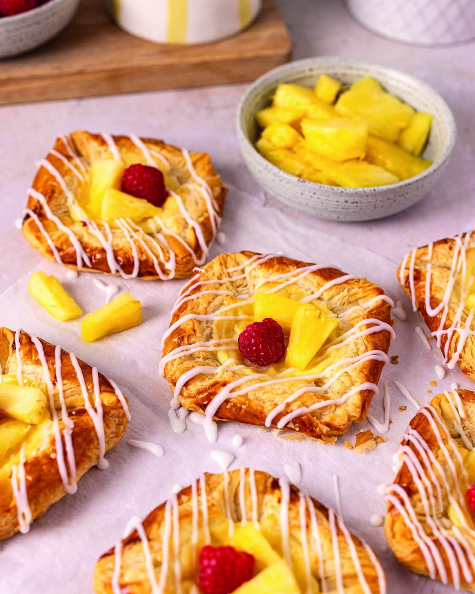 Frosted square pastries topped with raspberries and mango on a pastel table, with a bowl of mango chunks nearby