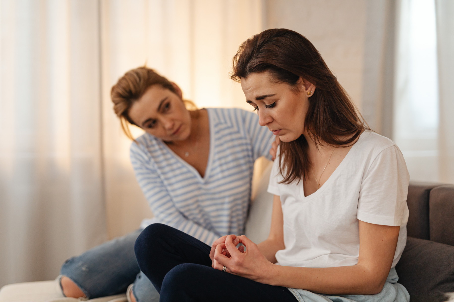 Woman sitting on a couch, looking upset while another woman leans in supportively beside her.