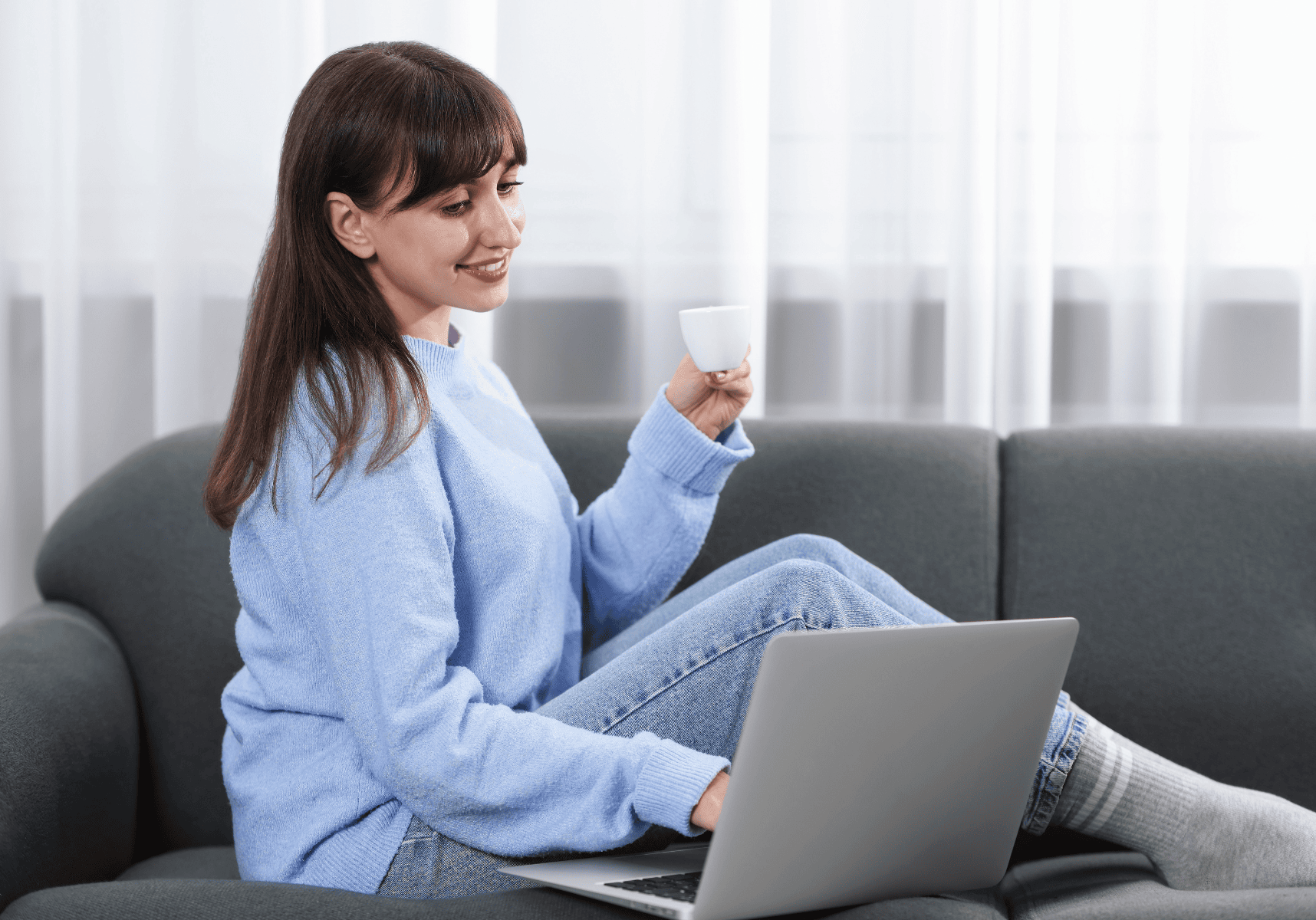 Woman on a gray sofa using a laptop while holding a cup, smiling in a bright room