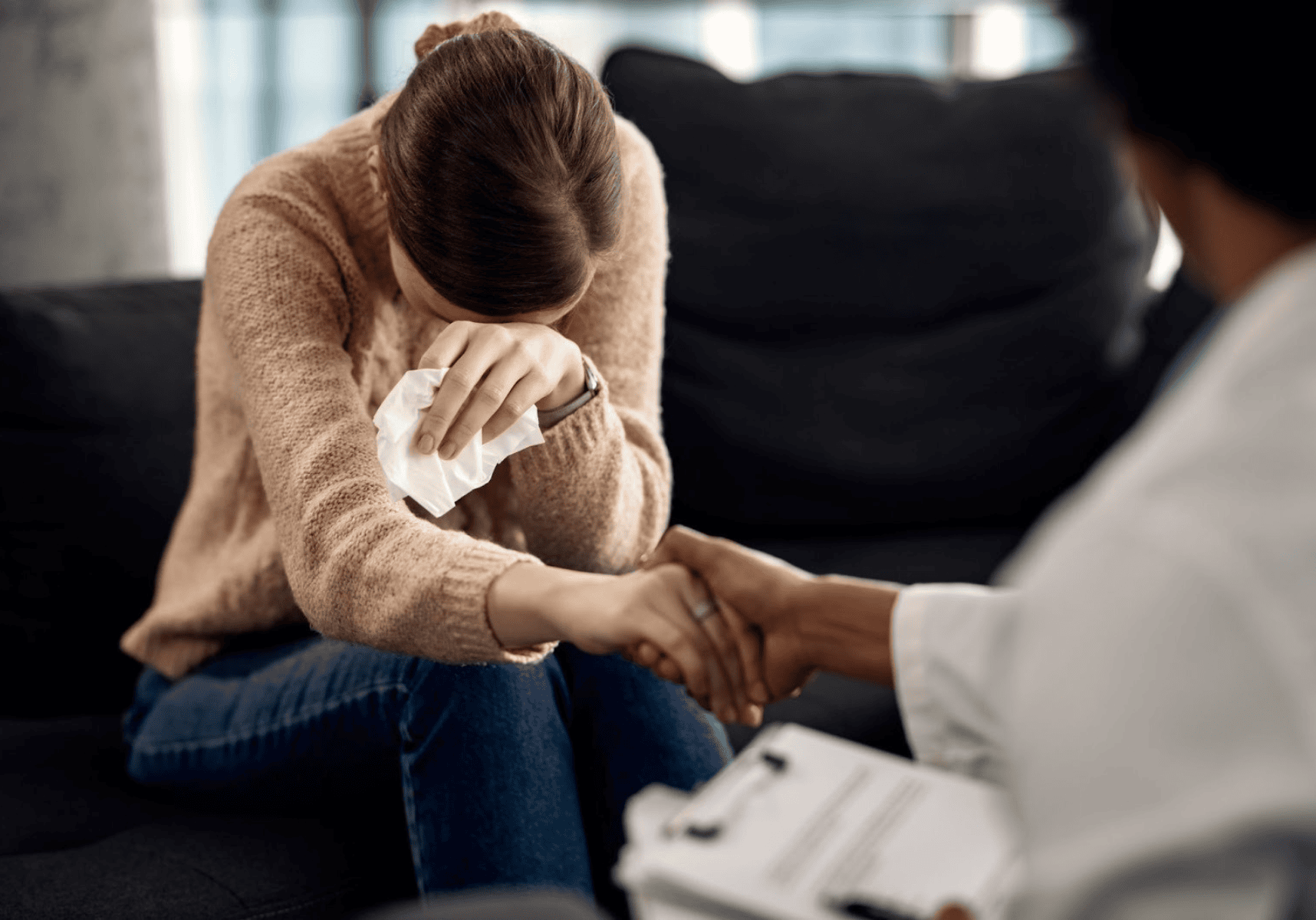 Person sitting with head in hands while another offers support in a counseling setting