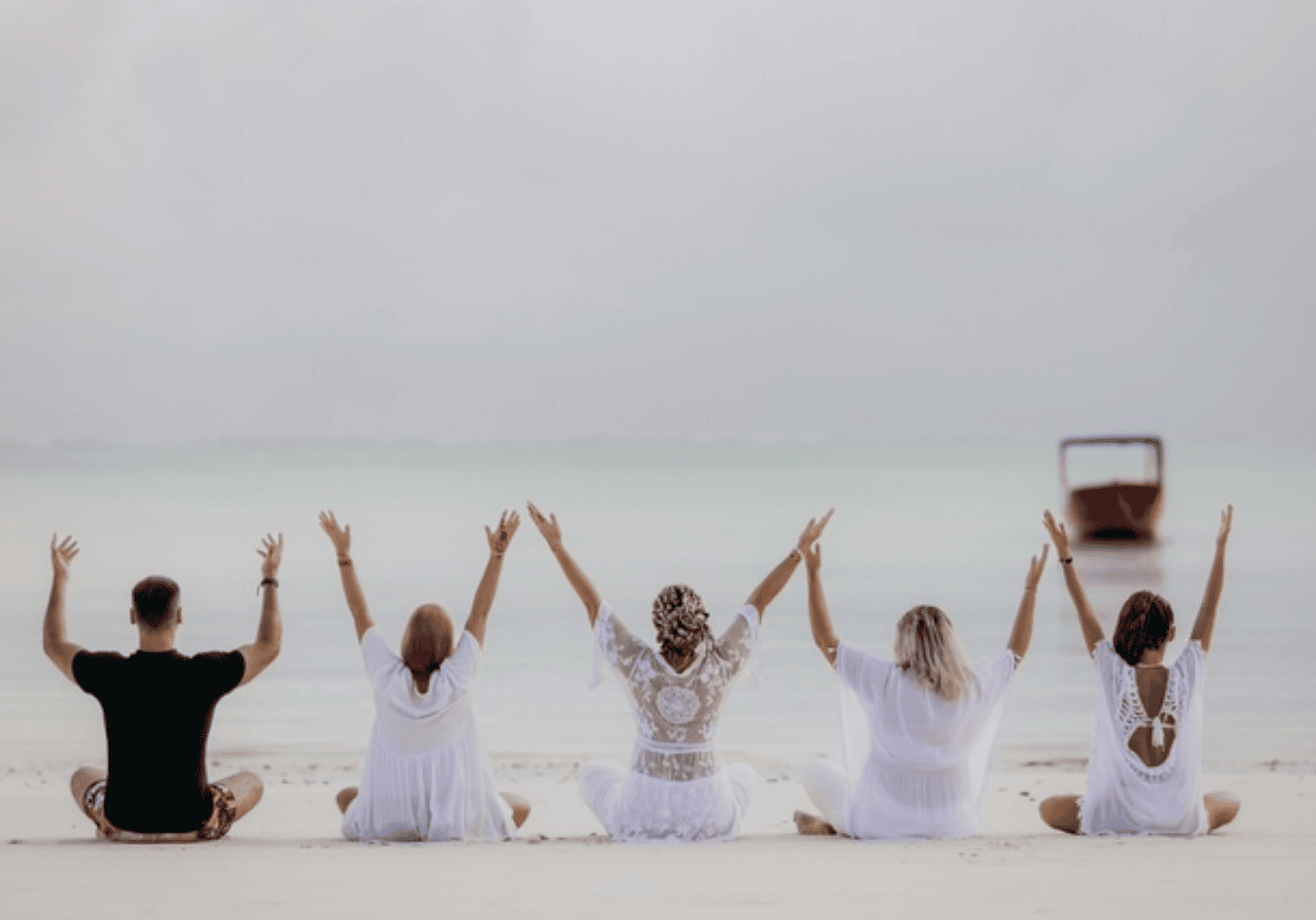 Five people sitting on a beach with arms raised toward the sea at sunrise