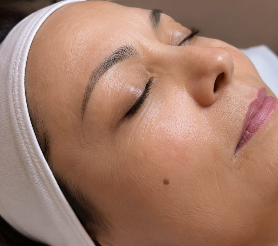 Primo piano di una persona con gli occhi chiusi, che indossa una fascia bianca per capelli, mentre si rilassa durante un trattamento viso.