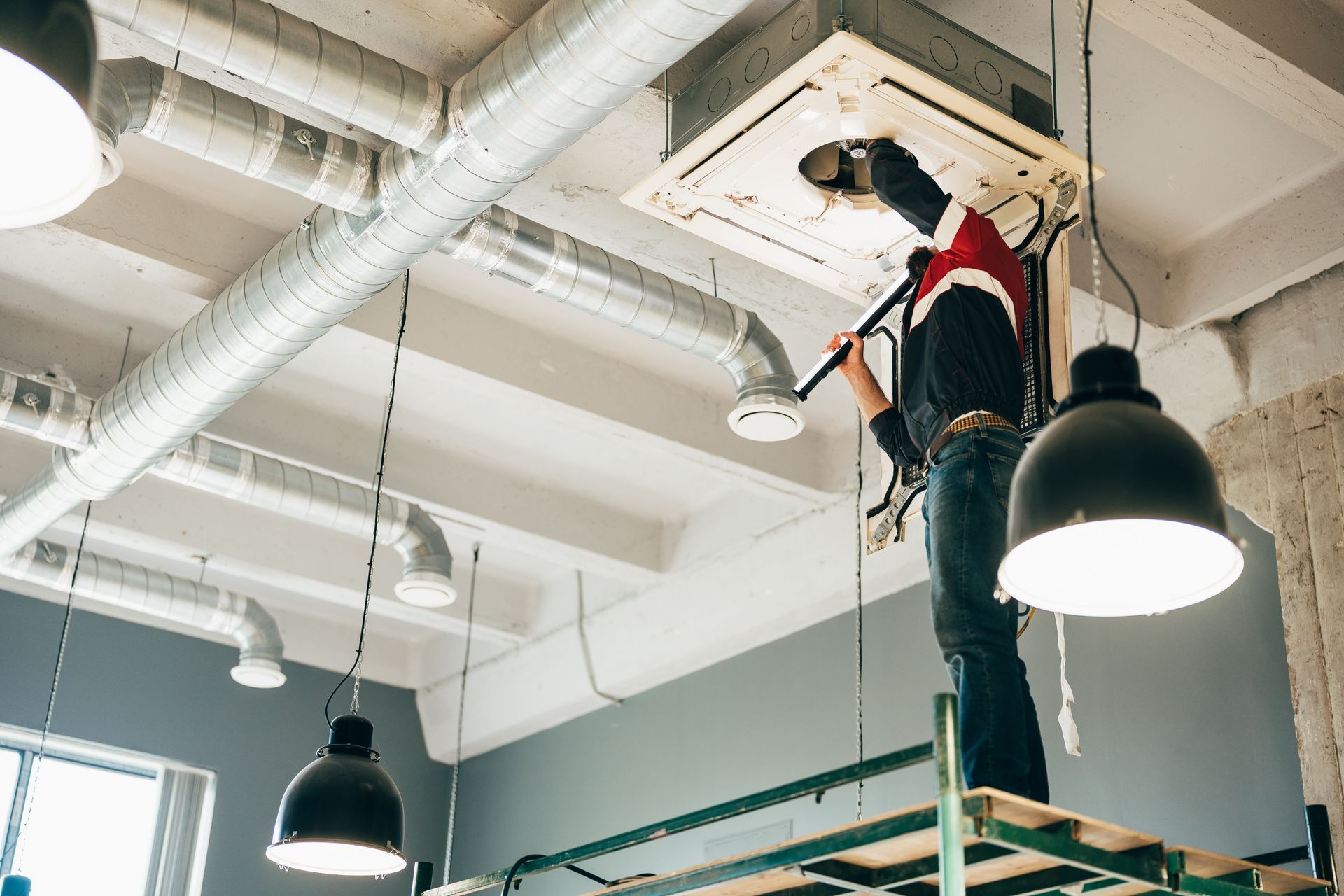 A technician servicing a ceiling-mounted commercial HVAC cassette unit with exposed ductwork above