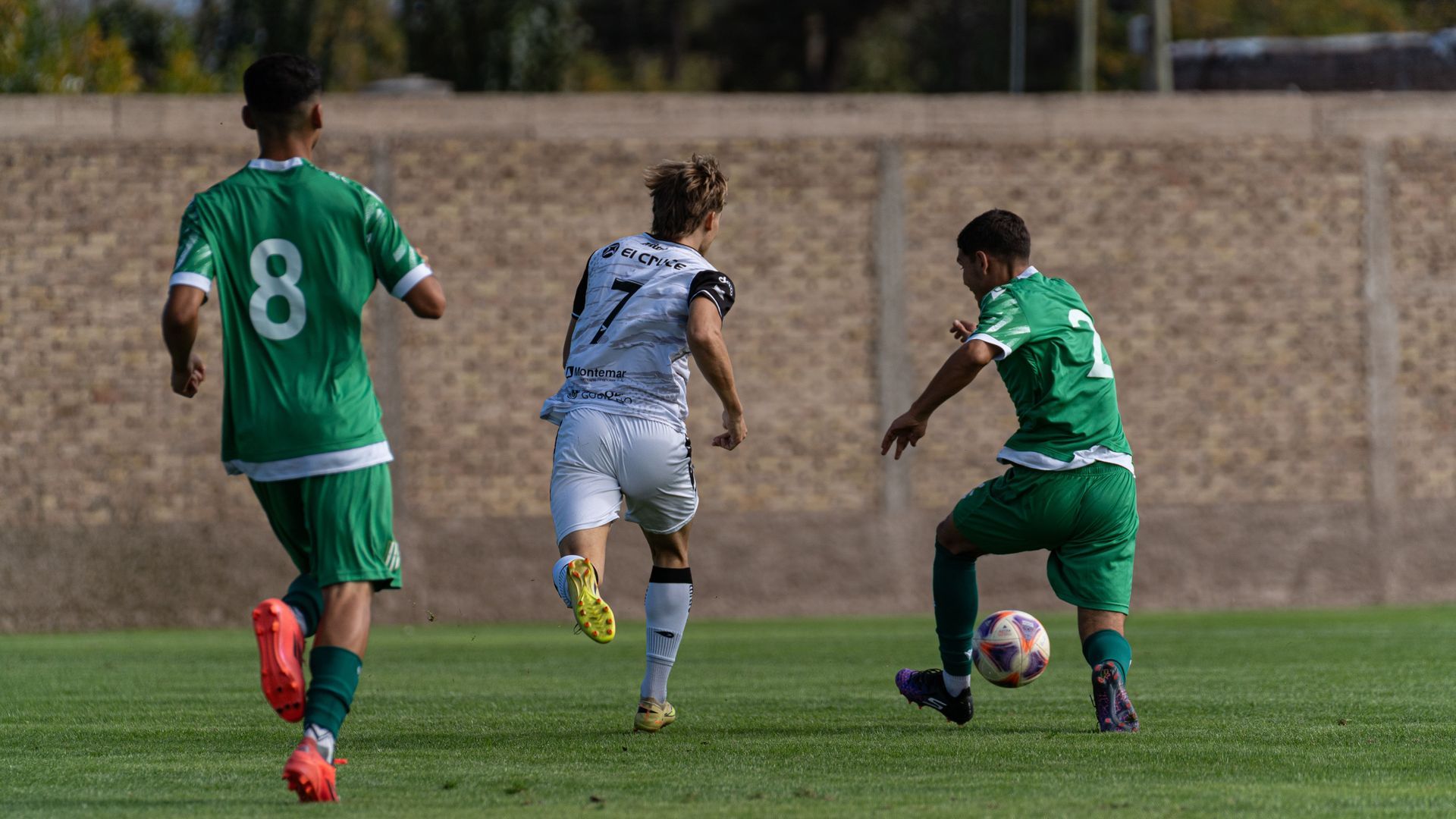 Three soccer players chase the ball on a grassy field, with two in green and one in gray.