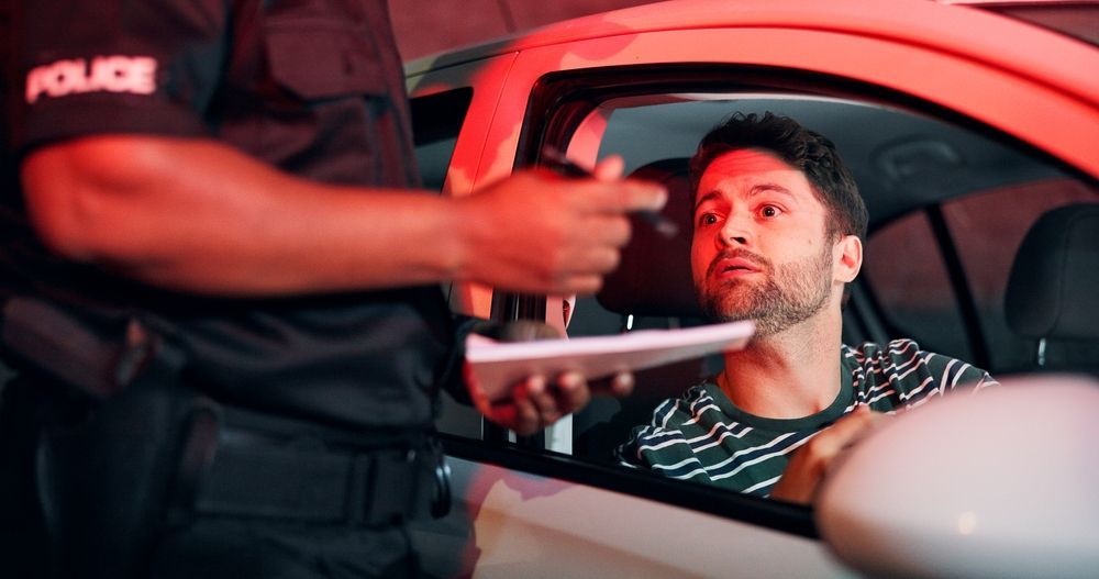Police officer at car window, pointing. Driver inside car looks concerned under red emergency lights.