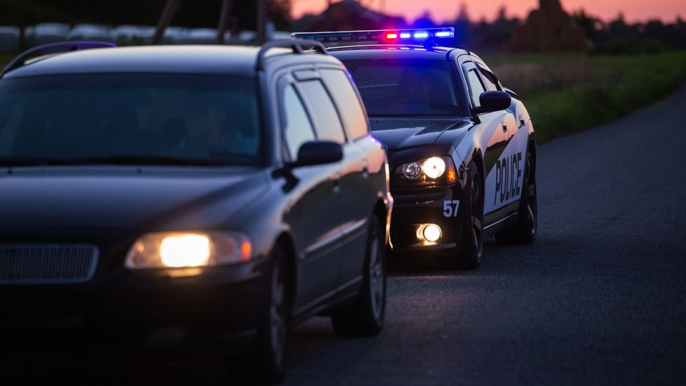 Police car with flashing lights pulls over a dark sedan on a road at dusk.
