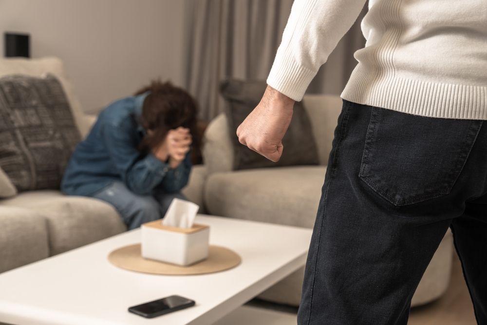 Man with clenched fist, facing woman on couch, hands covering face. Tissue box and phone on coffee table.