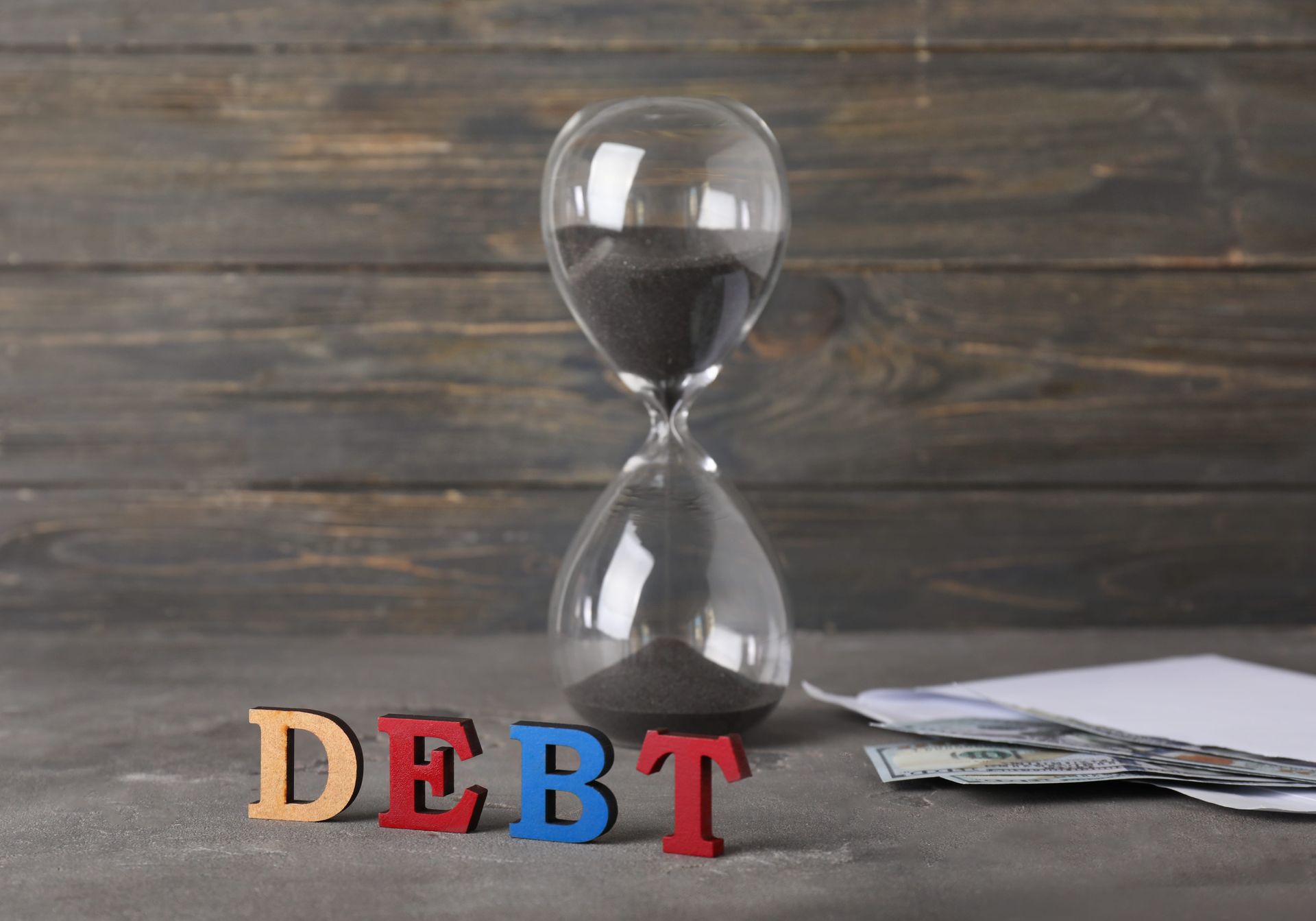 Hourglass, colorful DEBT letters, and scattered bills on a wooden surface