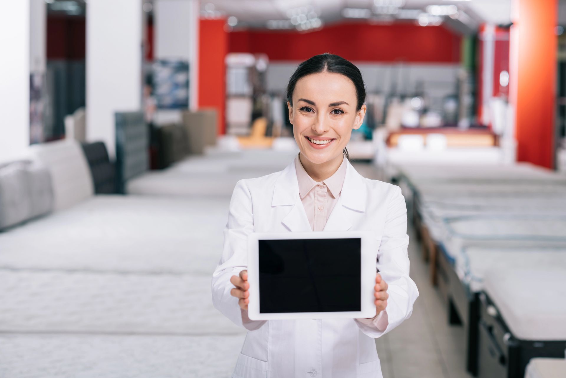 Un professionista sorridente in camice bianco è in piedi in un negozio di materassi, con in mano un tablet dallo schermo vuoto.