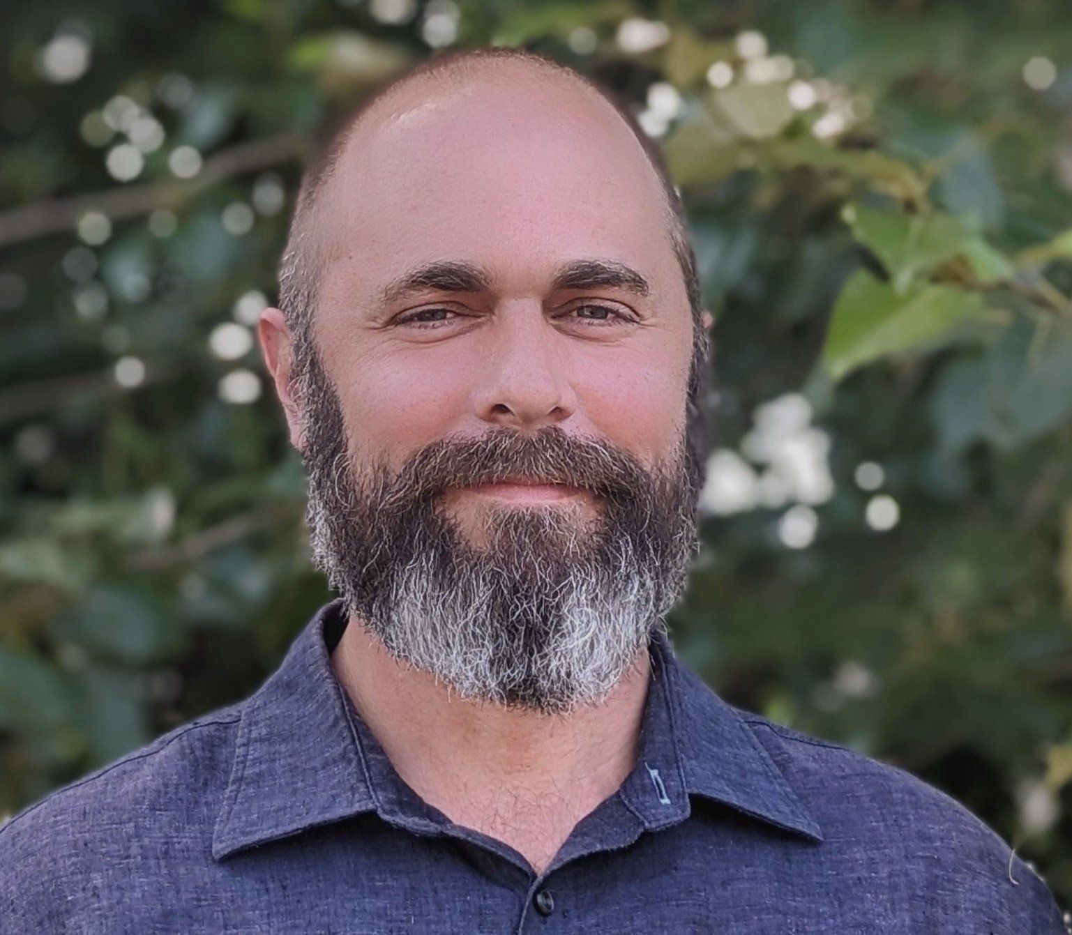 Man with a beard and short hair smiles, wearing a blue shirt, outdoors in front of leafy green background. Man with a beard and short hair smiles, wearing a blue shirt, outdoors in front of leafy green background.