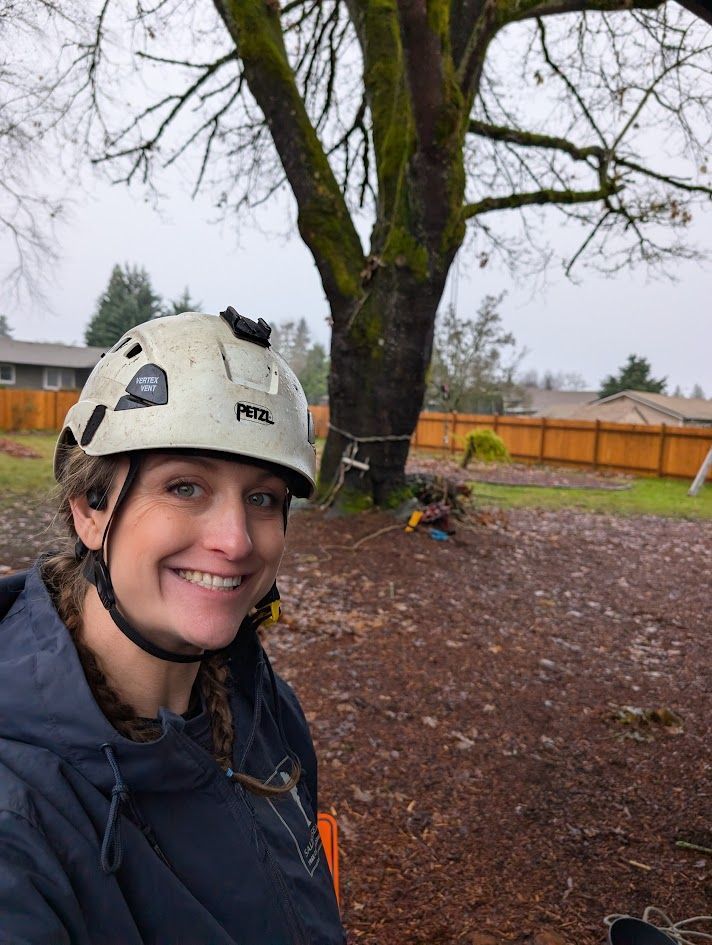Woman with helmet smiles in front of a tree, likely an arborist. Outdoors with brown ground and fence. Woman with helmet smiles in front of a tree, likely an arborist. Outdoors with brown ground and fence.
