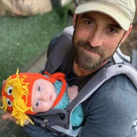 Man holding baby in a carrier, both looking at the camera. Baby wears a monster hat. Man holding baby in a carrier, both looking at the camera. Baby wears a monster hat.