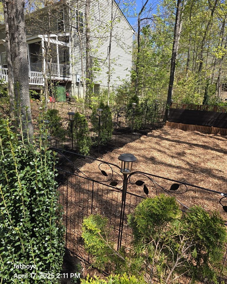 Wooded backyard with black fence, shrubs, mulch, and a house partially visible on the left.