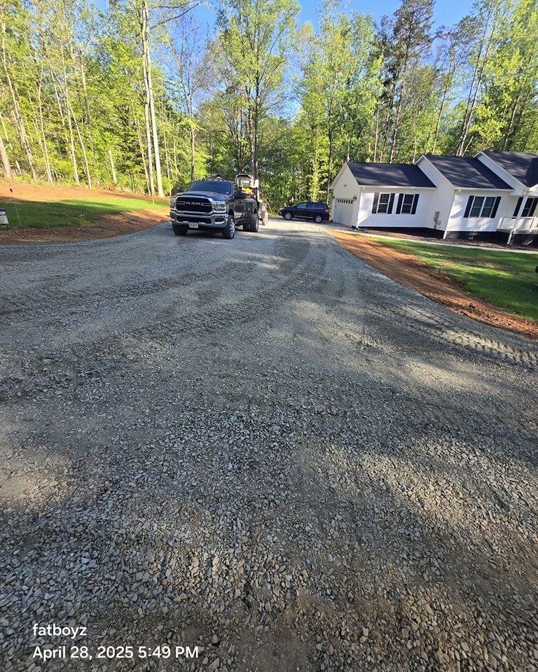 Gravel road under construction with trucks parked near a white house and trees along the roadside.