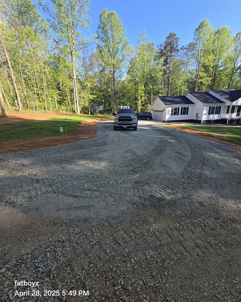 Gravel driveway with a parked black SUV beside a white house, surrounded by trees under a clear blue sky