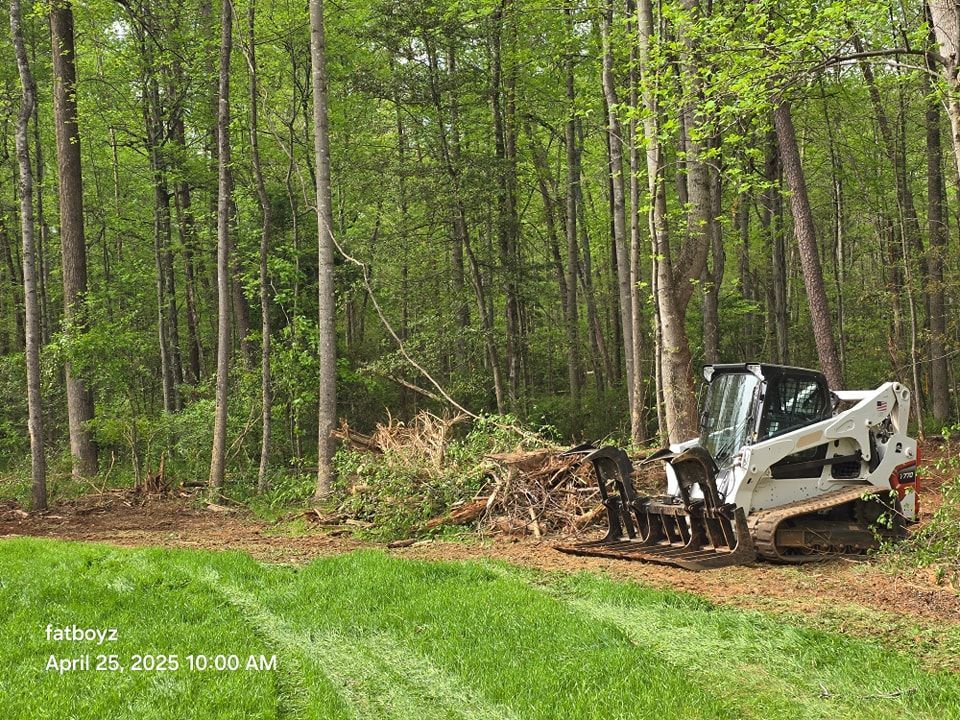 White skid steer beside a brush pile at the edge of a green field in a wooded area.