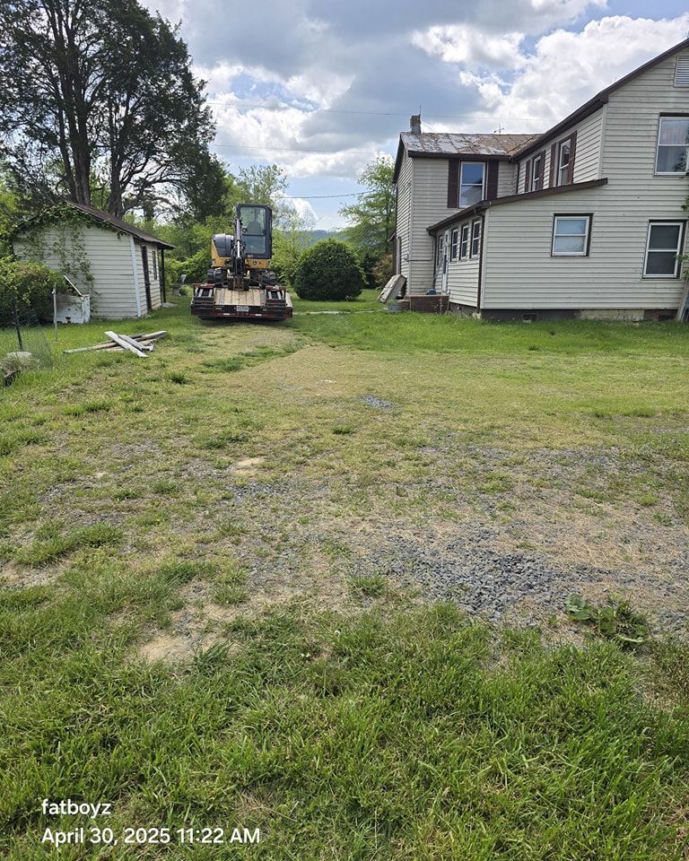 Yellow excavator in a grassy yard beside a white house and small shed under a cloudy sky