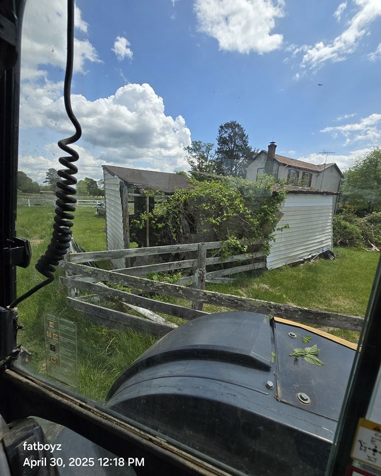 Rural yard with weathered fence, white shed, and cloudy blue sky, viewed from inside a vehicle.