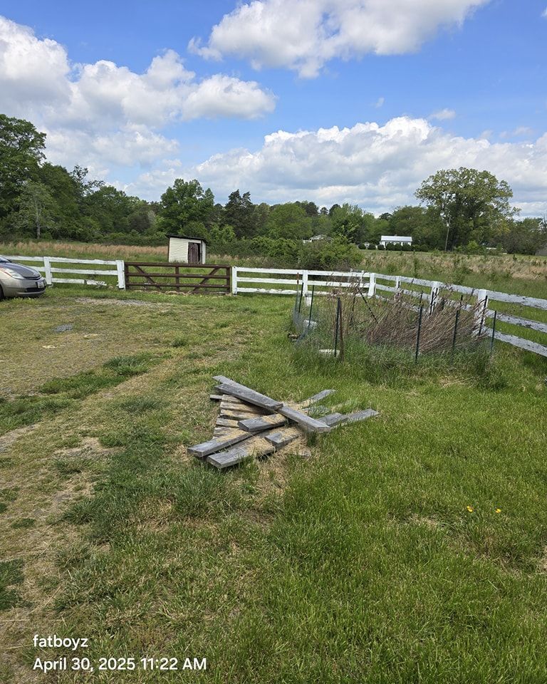 Rural fenced yard with white fence, grass, trees, and scattered wooden boards under a cloudy blue sky