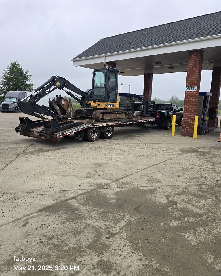 Excavator loaded on a flatbed trailer under a gas station canopy on a wet concrete lot.