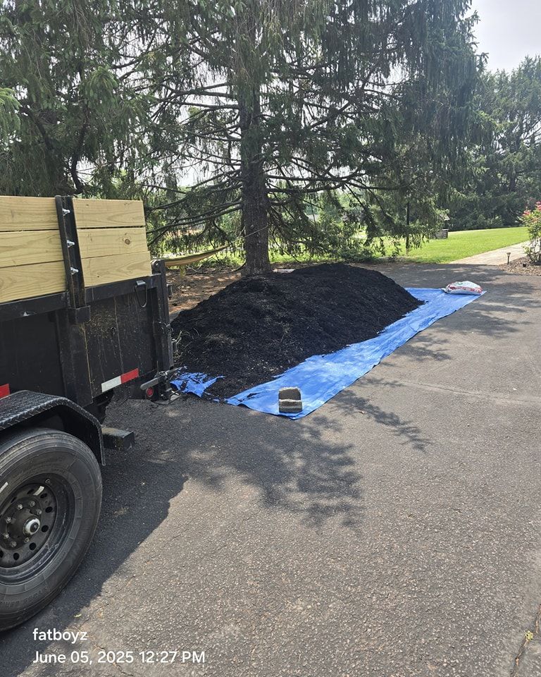 Black mulch pile on a blue tarp beside a trailer on a paved driveway under trees.