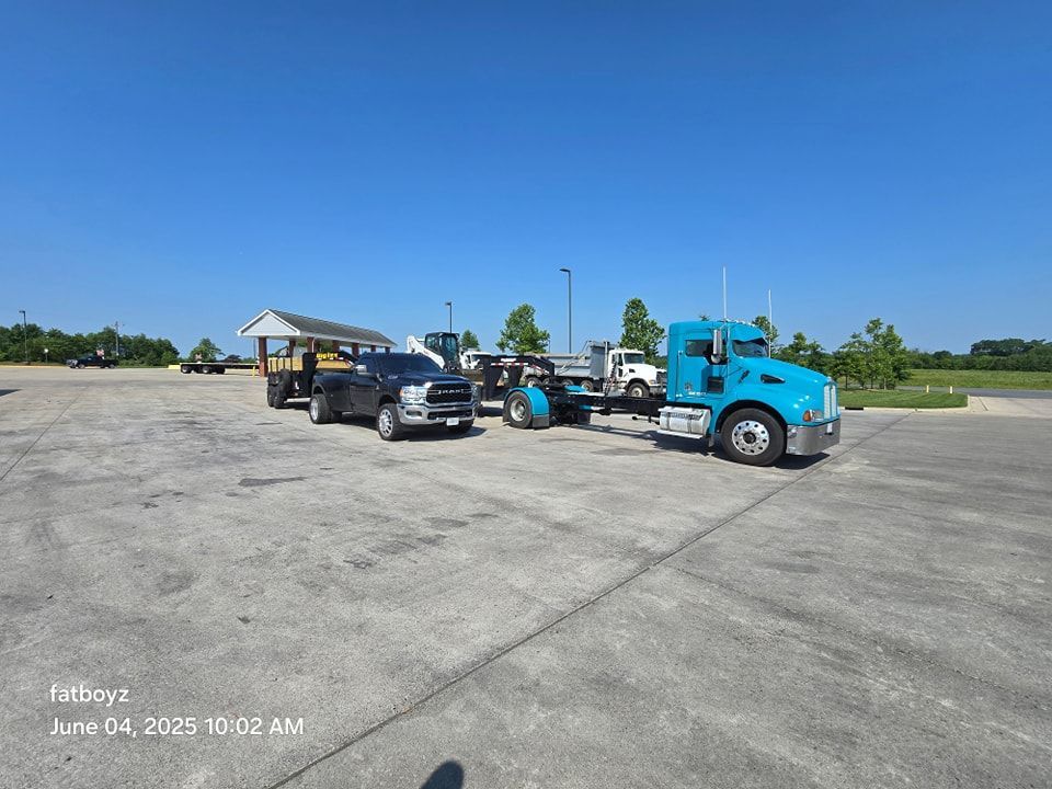 Blue semi truck with a flatbed trailer and black pickup parked on a wide open lot under a clear sky
