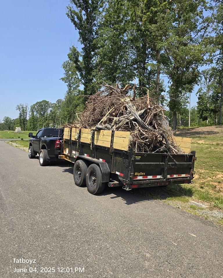 Black pickup towing a trailer loaded with tree branches along a rural road under a blue sky