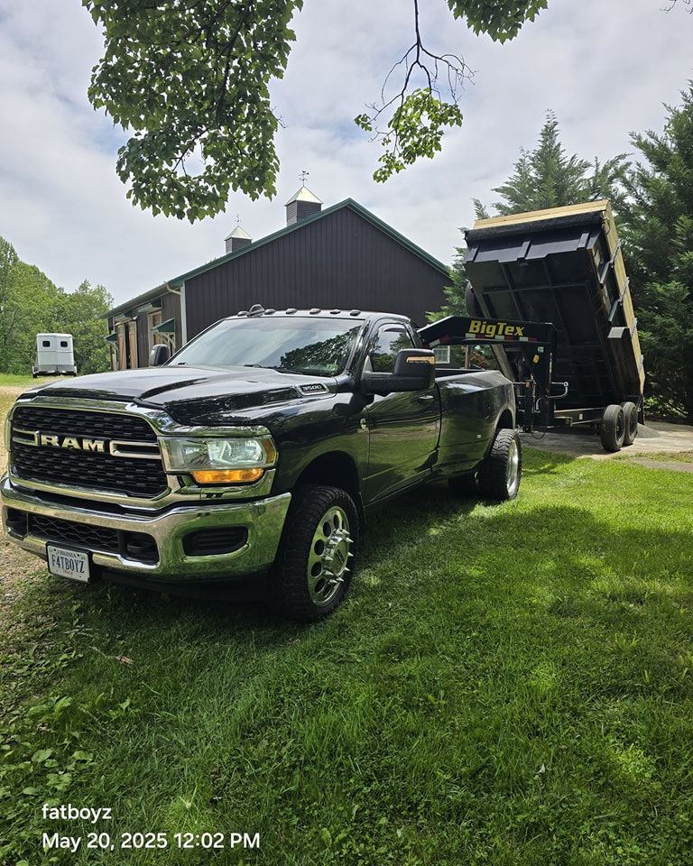 Black RAM truck parked on grass beside a house, unloading debris from a raised dump trailer.