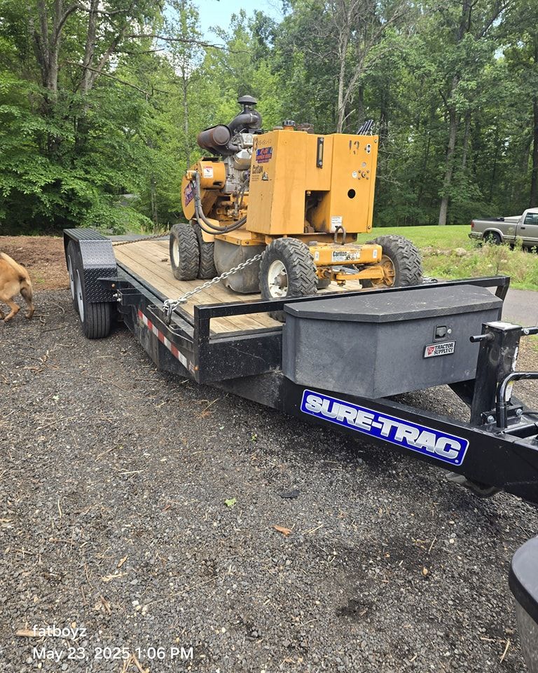 Yellow skid-steer loader on a flatbed trailer in a wooded gravel lot.