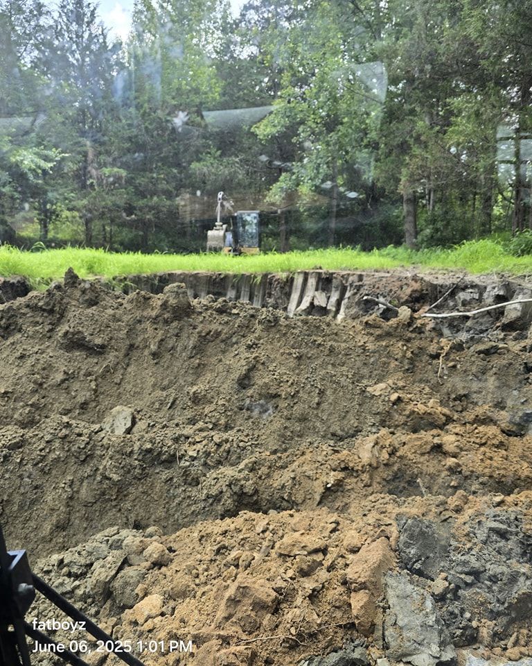Large muddy excavation pit beside a grassy area with trees and a small structure in the background