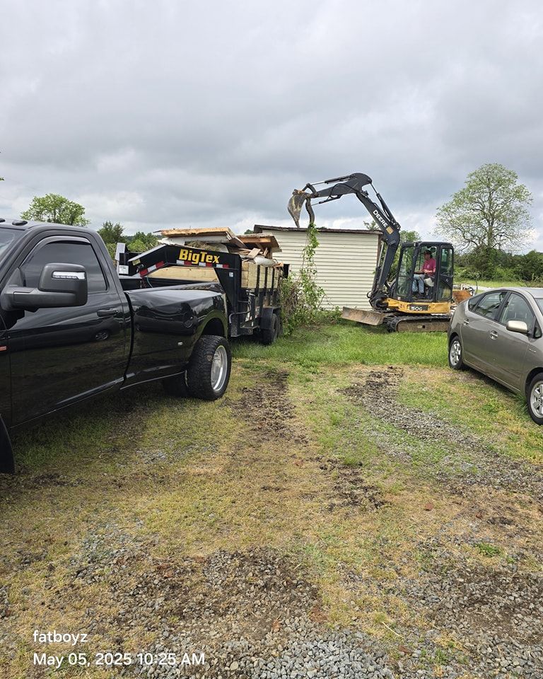 Excavator and trailer beside a white shed, with black pickup trucks on a muddy yard under cloudy skies