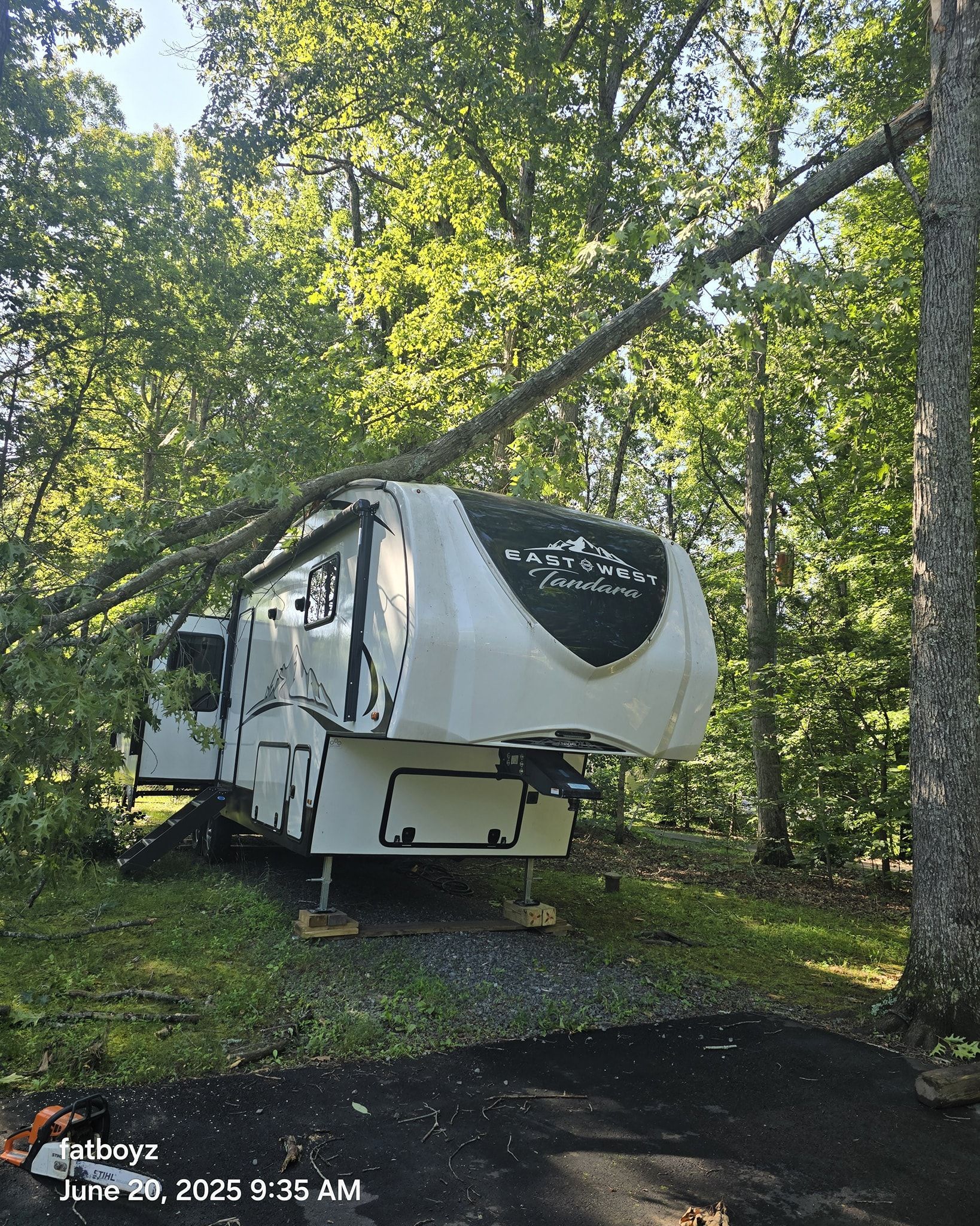 White RV trailer parked among trees in a wooded campground, with a paved site in front.