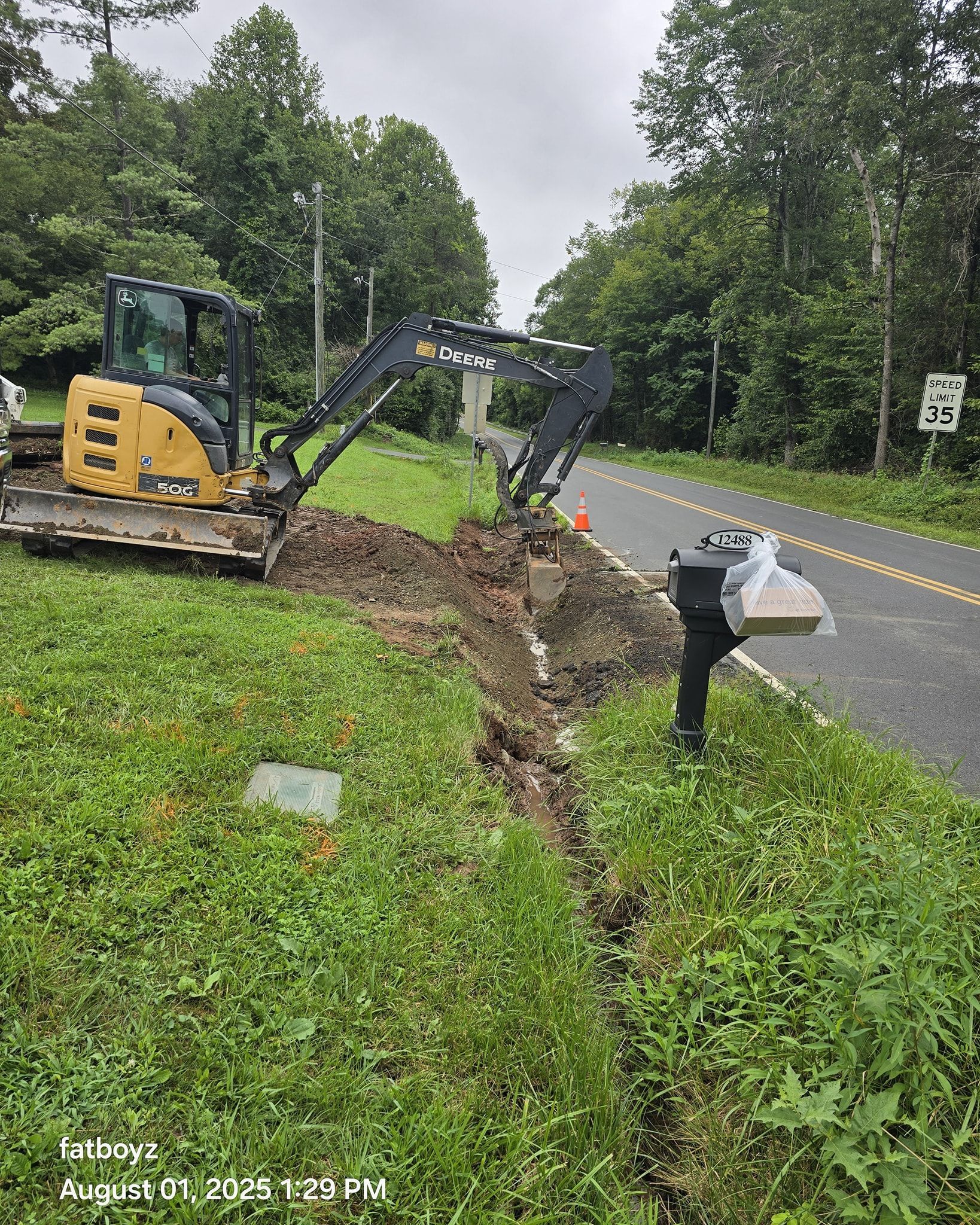 Construction equipment digging a trench beside a wet road in a wooded area