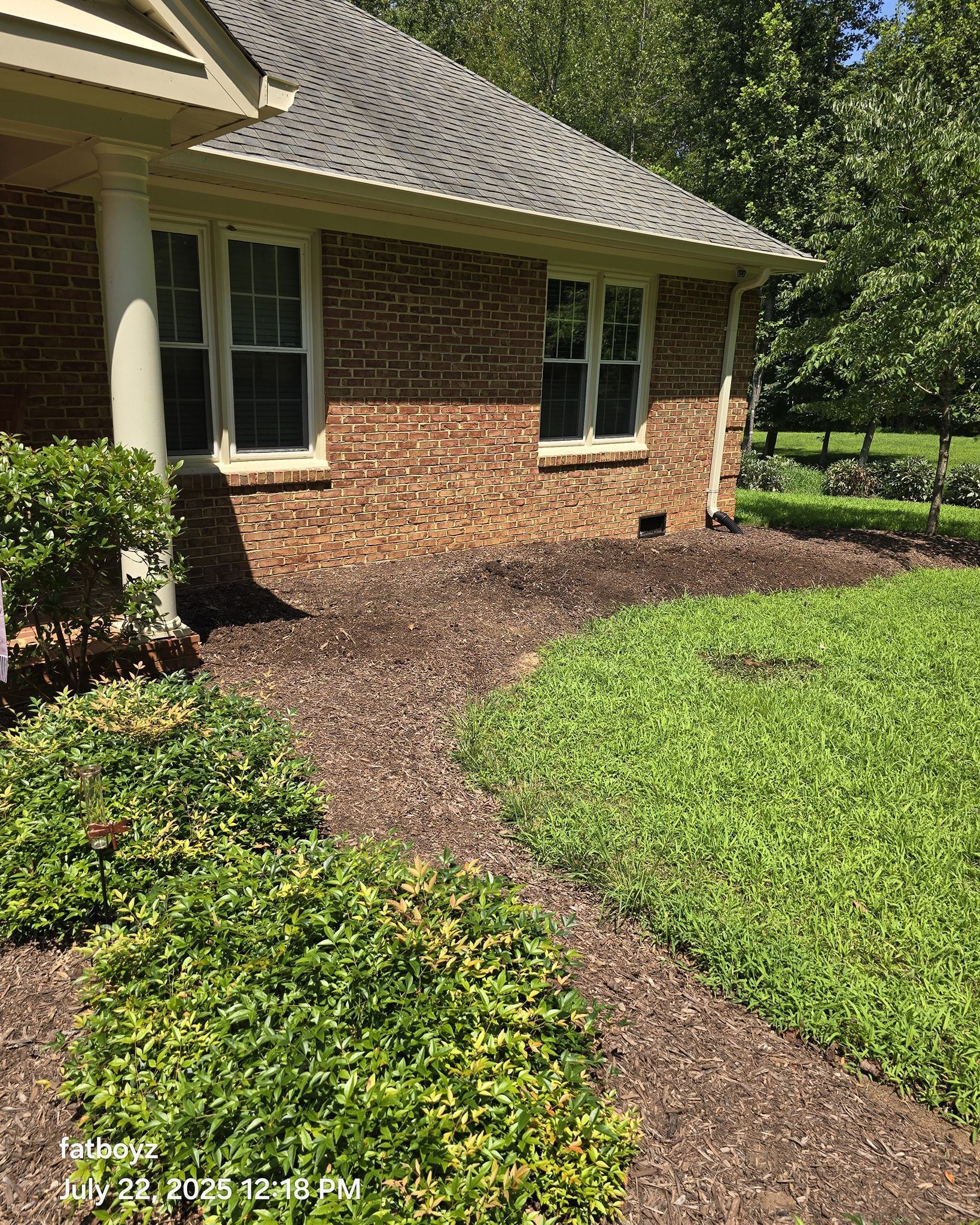 Brick house exterior with white columns, trimmed shrubs, and a mulch path in bright sunlight