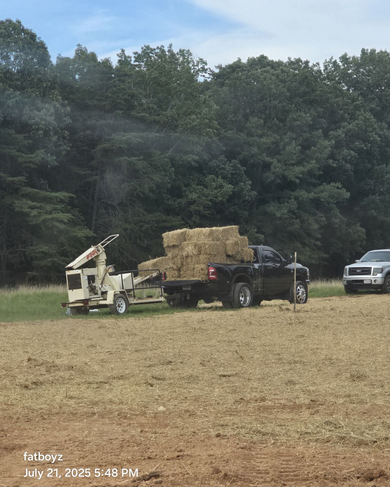 Hay wagon loaded with hay bales in a field, with trucks and a wooded tree line behind.